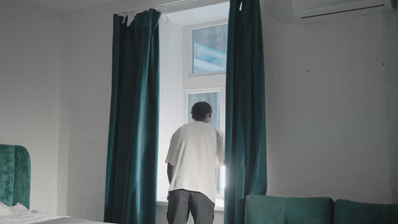 Man in white t-shirt pulling back dark green curtains in a minimalist bedroom, allowing natural light to fill the room, with neatly made bed, green headboard, and decorative window shutter