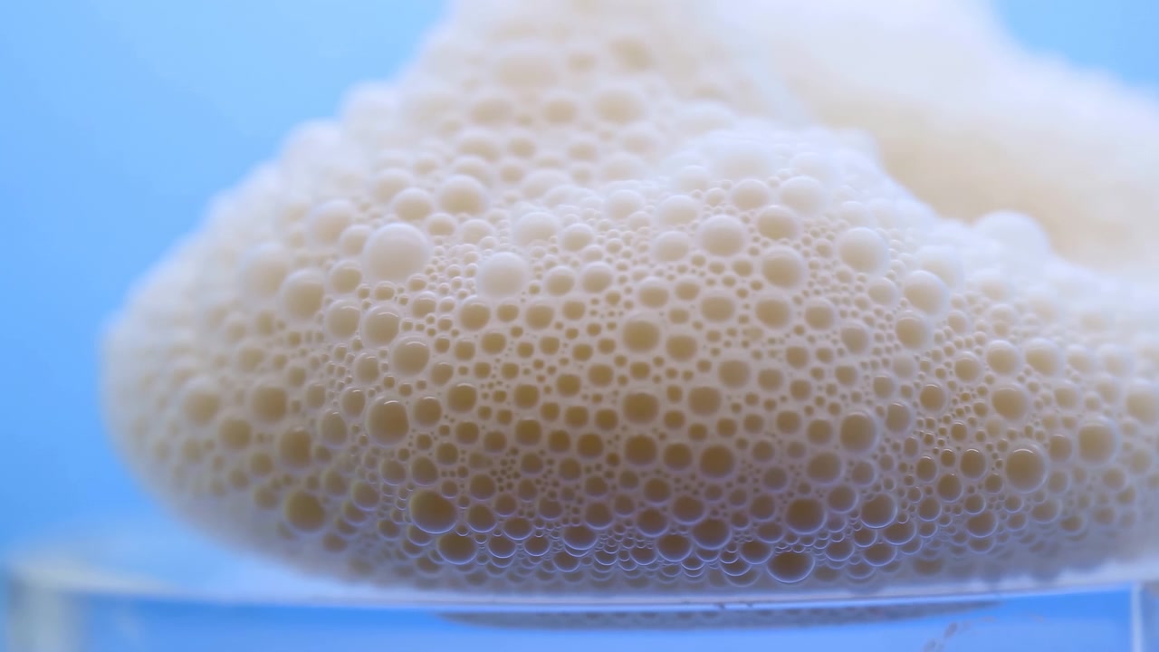 Vista baja de espuma de crema espesa en el borde de un vaso transparente, mostrando burbujas densamente agrupadas de varios tamaños, reflejos suaves, textura lisa, tonos pálidos y un fondo azul frío, con espuma y burbujas.