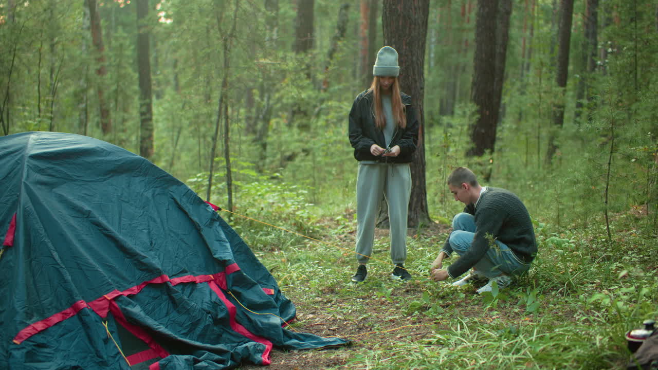 Man squatting on forest floor receives tent peg from lady in casual outfit, hammers it into ground beside green and red tent surrounded by trees, preparing campsite