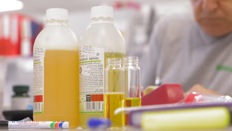 Laboratory-table-full-of-test-tubes-and-beakers-with-yellow-liquids-and-a-blurred-person-in-the-background-working---trucking-to-right