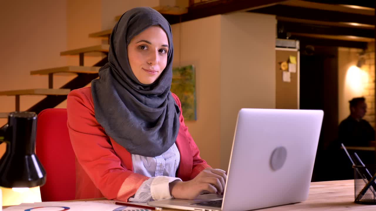 히자브를 입은 젊은 아름다운 무슬림 사업가 여성이 노트북으로 타이핑하고 카메라를 보고 행복하게 미소 짓는 클로즈업 초상화