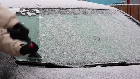 man in coat and gloves cleaning frozen car windshield, scrapes ice off the glass surface in winter