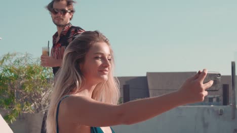 young man holding a drink and taking a selfie with a blonde young woman sitting in a hammock in a resort