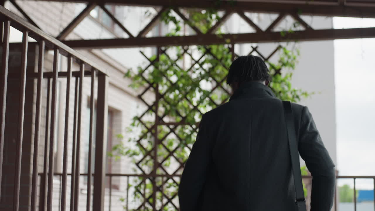 두꺼운 정장을 입고 숄더백을 메고 야외 복도를 걷는 학생의 뒷모습이에요. 금속 난간과 녹색 덩굴이 격자 모양으로 산책로를 둘러싸고 있어 도심 캠퍼스 분위기를 연출해요.