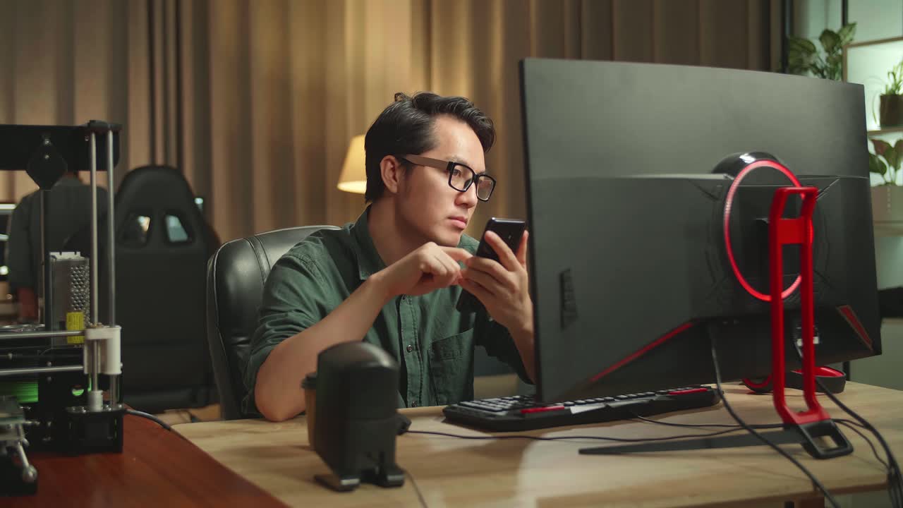 Asian Man Using Mobile Phone While Works On Personal Computer And 3D Printer In Home Office, He Works At Night