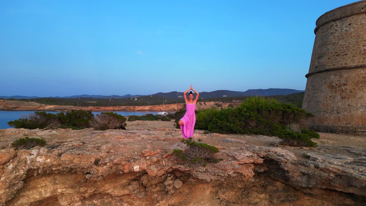 Mulher despreocupada de vestido rosa com braços abertos desfrutando da liberdade e de uma vista deslumbrante na ilha de Ibiza, perto da Torre des Savinar ao pôr do sol. Drone ascendente.