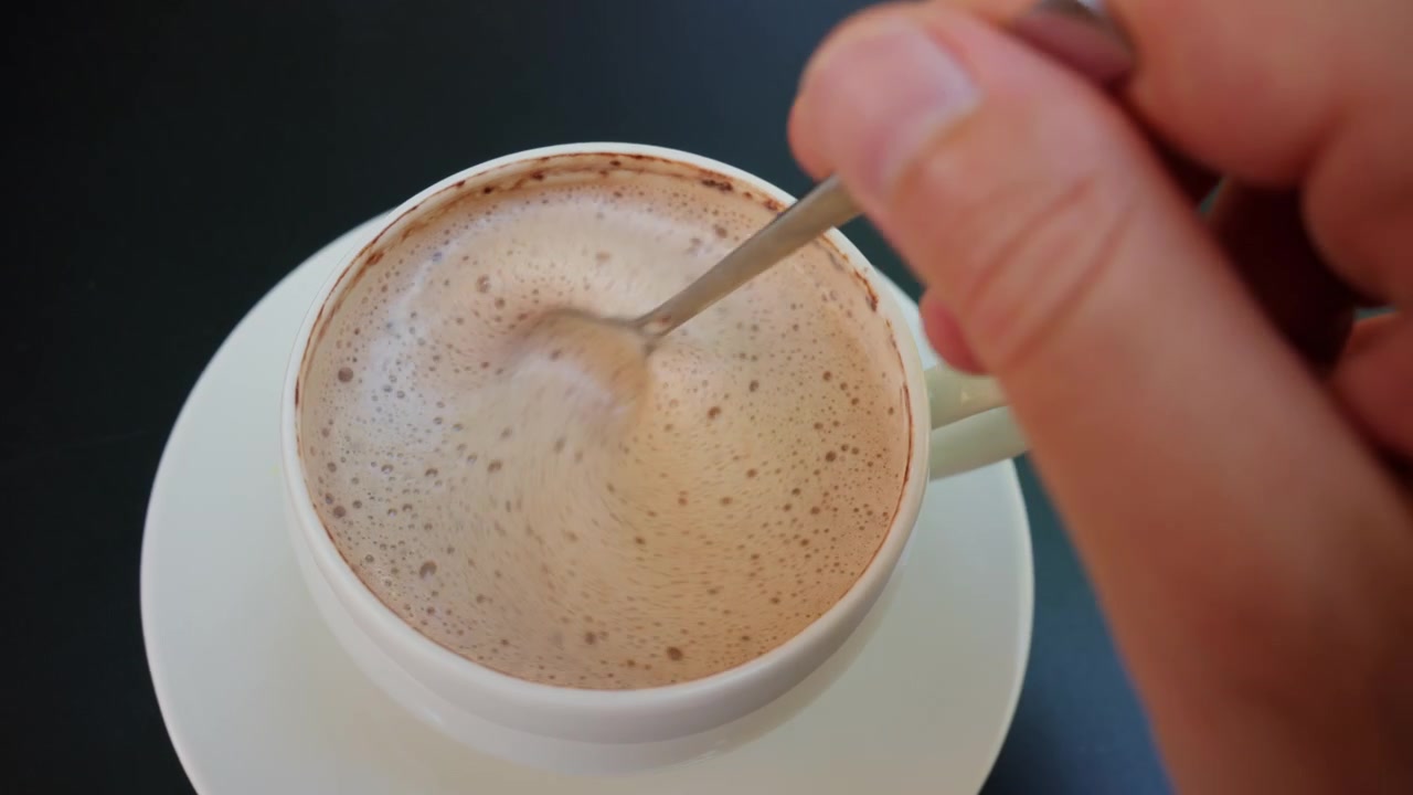 Persona che mescola caffè caldo o latte macchiato con un cucchiaino in una tazza e piattino bianchi, mostrando la schiuma in superficie.