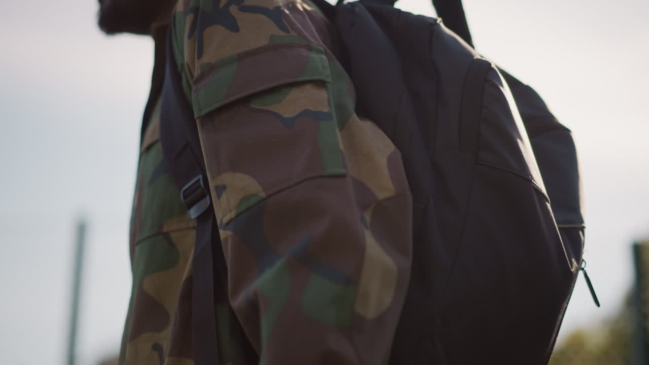 Soldier With Backpack, Person In Camouflage Jacket Moves Purposefully Near Fence At Sunset, Individual Equipped With Pack And Camouflage Clothing Advances Through City Training Zone Peacefully