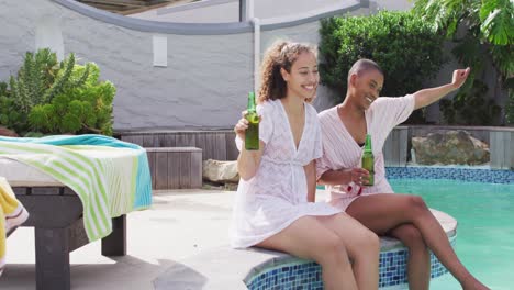 Two-happy-diverse-female-friends-sitting-with-beer-and-having-fun-at-pool-party