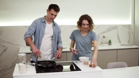 casual couple cooking together in domestic kitchen. positive pretty woman breaking eggs and handsome husband cooking omelet pouring oil on hot pot