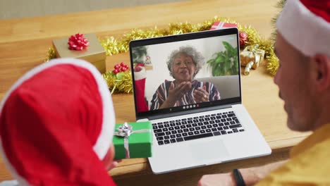 padre caucásico con hijo con sombreros de santa usando una computadora portátil para una videollamada de navidad, con el hombre en la pantalla