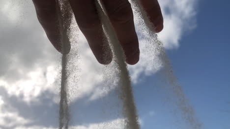 Cámara-Lenta-De-Granos-De-Arena-Cayendo-En-Un-Concepto-De-Cascada-De-Una-Mano-Con-Un-Fondo-De-Cielo-Azul,-Símbolo-Del-Tiempo-Que-Pasa