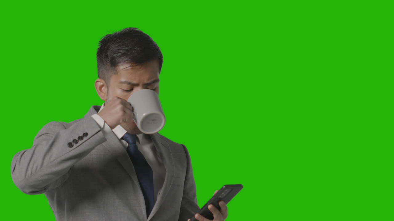 Scatto in studio di un uomo d'affari in abito che tiene una bevanda calda mentre guarda il cellulare su sfondo verde