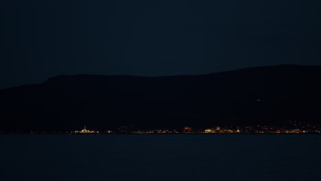 coastal town lights up at night with ferry boats crossing paths in front below mountains