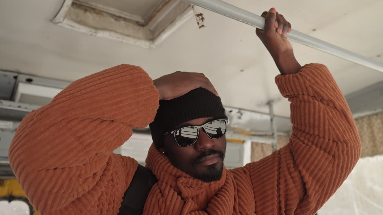 Uomo di colore con maglione arancione che afferra il corrimano, ritratto di pendolare all'interno di un autobus urbano, berretto di lana e occhiali da sole, tracolla dello zaino visibile, strati invernali e texture accogliente, finestra riflettente con sfocatura della città, morbido.