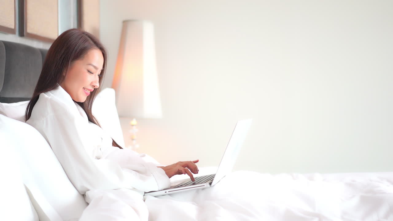 호텔 객실 침대에 누워 있는 노트북 컴퓨터를 사용하여 흰색 목욕 가운을 입은 아시아 여성의 옆모습을 보고 웃고 있는 노트북 키보드에 메시지를 입력하고 있습니다.