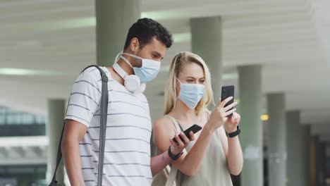 Side-view-of-Caucasian-couple-on-the-go-wearing-a-coronavirus-covid19-mask-and-using-phone