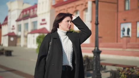 russian woman in black coat and white sweater adjusts her hair while carrying a black handbag and looking left, with blurred residential buildings in the background