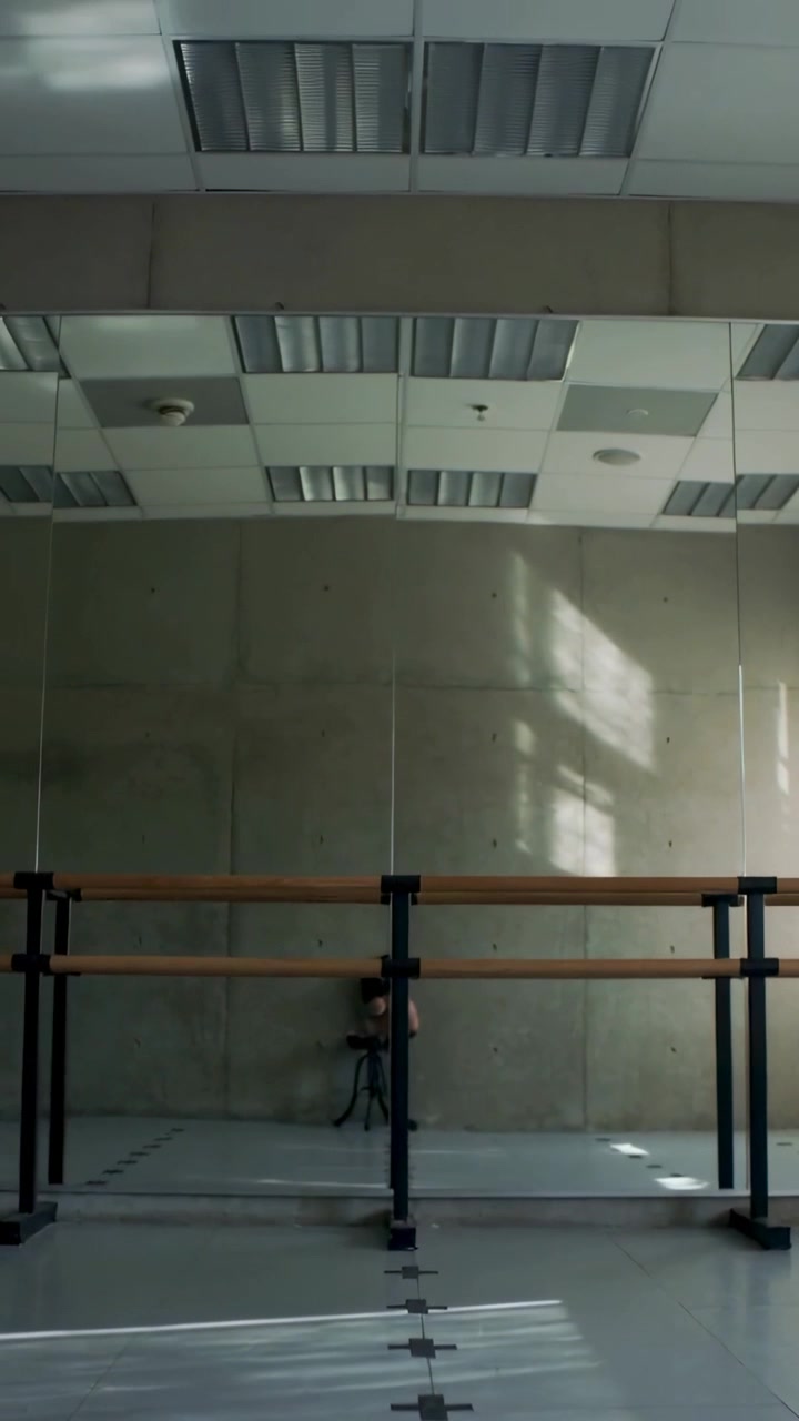 Vertical video: Recording dancer in dark clothes adjusting posture in rehearsal with barre, tripod