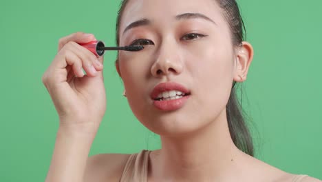 close up de una mujer asiática peinando sus largas pestañas negras con un pincel de máscara y sonriendo mientras está de pie en el fondo de la pantalla verde en el estudio