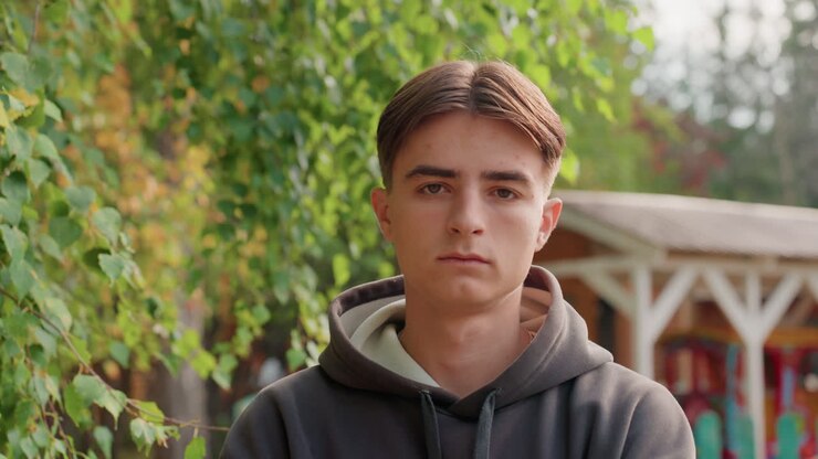 White teenage boy hoodie standing stoic against leafy backdrop and wooden fence. Reserved athlete folding stance in defiant pose, introspective poet rehearsing lines, streetwear model posing