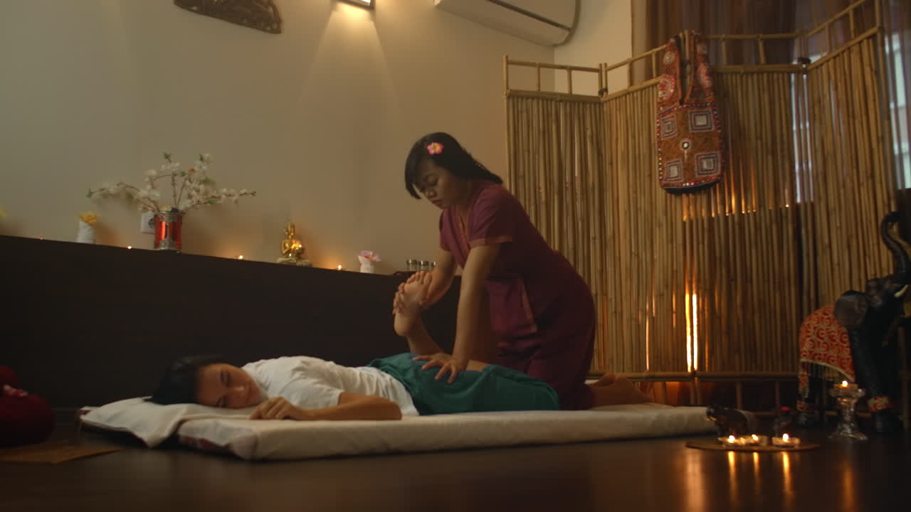 Asian Woman performs Traditional Thai Massage to beautiful European Woman. Rehabilitation and Treatment after Injuries with the help of Massage. Relax and Rest from massage of Legs Arms and Back. Therapeutic massage