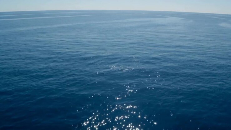 Vista alta e centrata dell'oceano aperto con acqua calma e blu profondo, leggere increspature vicino al primo piano, bande lisce e stratificate sulla superficie, sottile scintillio della luce solare e un orizzonte lontano e chiaro