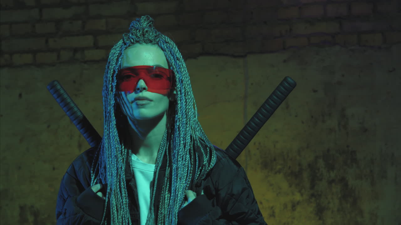 chica con dreadlocks y katanas en gafas rojas posando contra una pared de ladrillo de neón
