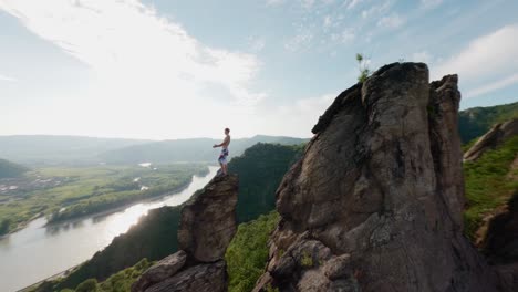 guy-on-top-of-mountain-ridge,-fast-drone-FPV,-speed-aerial-shot,-king-of-the-world-pose,-mountainclimber,-person-on-top-of-hill,-danube-river-valley-landscape-overview,-flying-around,-flying-fast