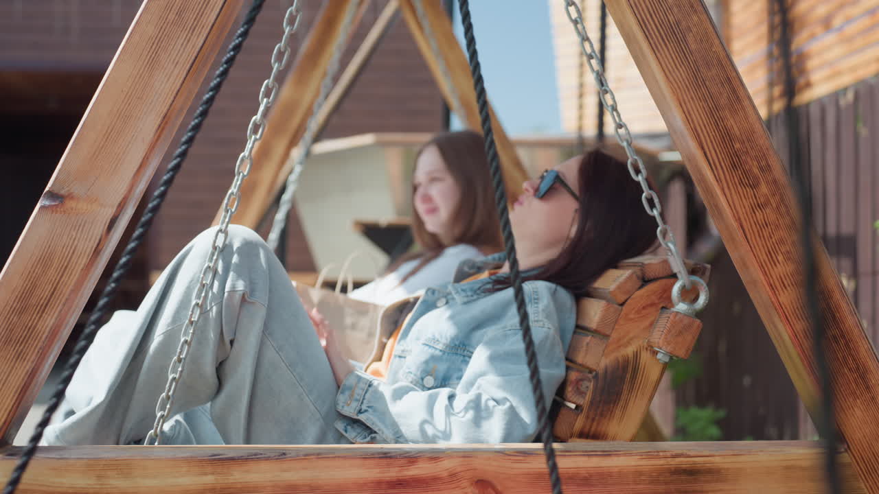 Two friends sit close together on wooden swing, gently swinging and enjoying warm sunlight, one wears denim jacket and sunglasses, both relaxed and calm in cozy outdoor backyard