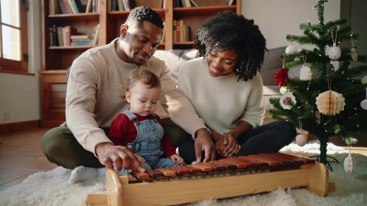 Il padre si allunga per premere le barre dello xilofono, mentre la madre guida il bambino piccolo sul tappeto vicino all'albero di Natale.