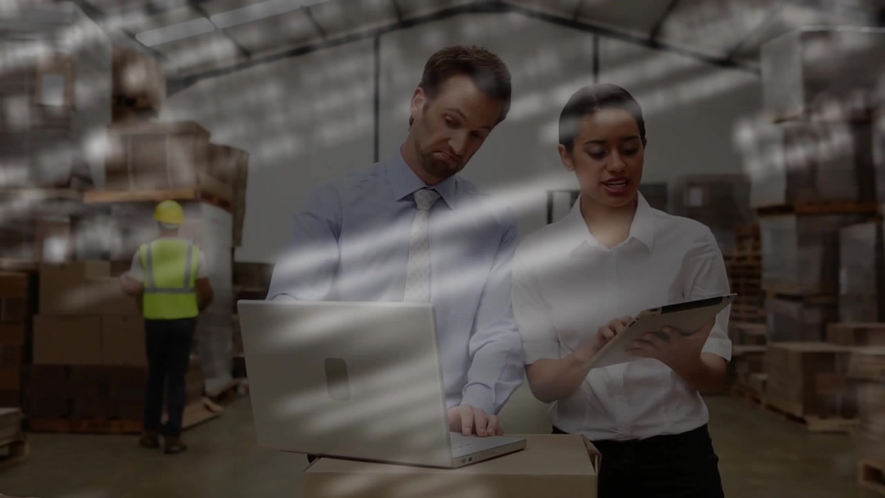 Responsabili della logistica che indicano un laptop e scorrono un tablet, monitorando i dati di inventario tra pile di scatole.