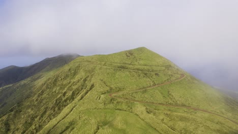 drone orbiting vulcanic mountain, pico da esperança, covered in lush green with low clouds in são jorge island, the azores, portugal