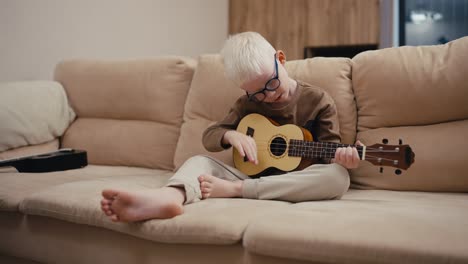 Little-albino-boy-with-white-hair-color-in-round-blue-glasses-plays-the-ukulele-while-sitting-on-a-cream-sofa-in-a-modern-apartment-in-the-evening.-A-little-albino-boy-with-white-hair-in-blue-glasses-sits-on-a-large-cream-sofa-at-home-and-plays-a-yellow-ukulele