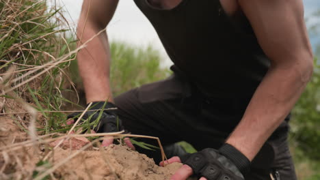 soldier rising from grass covered sand mound kicking up soil and sand with boot as loose pebbles scatter among wild stems and shrubs during remote outdoor trek beneath bright cloudy sky