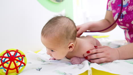 adorable baby lying naked on its belly and holding head up. pediatrician puts stethoscope to kid's back. close up.