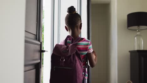 video of happy african american girl with schoolbag opening front door and leaving home for school