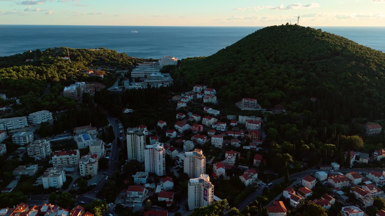 Aerial drone shot panning left over Dubrovnik at sunset, showing the historic town surrounded by lush green nature and calm blue Adriatic Sea under a glowing golden horizon with soft evening light