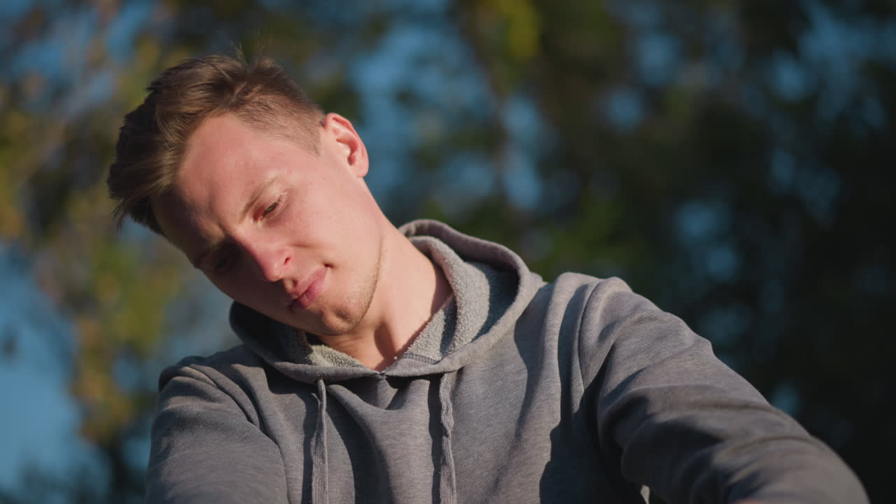 Close-up of young man in gray hoodie, focused on adjusting stilts in sunny outdoor with blurred greenery. His concentrated expression and careful grip on stilt handle are highlighted by warm sunlight