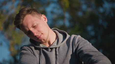 close-up of young man in gray hoodie, focused on adjusting stilts in sunny outdoor with blurred greenery. his concentrated expression and careful grip on stilt handle are highlighted by warm sunlight