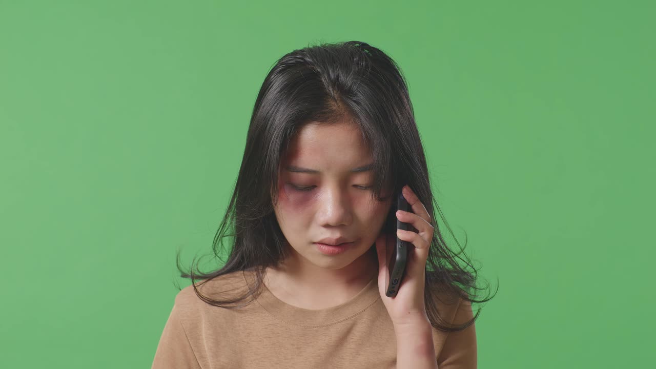 얼굴에 이 있는 젊은 아시아 여성의 폭력 피해자가 녹색 스크린 배경 스튜디오에서 스마트폰으로 도움을 요청하는 동안 울고 있습니다.