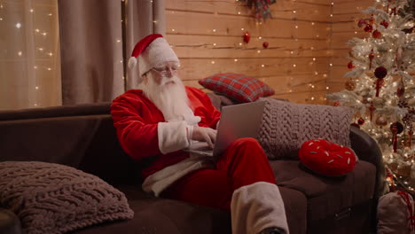 santa relaxes at home near a decorated christmas tree with a laptop and answers messages. santa claus on the eve of birth at home