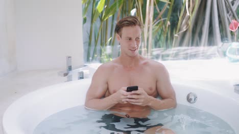 young man uses smartphone sitting in modern round bathtub at luxury apartments