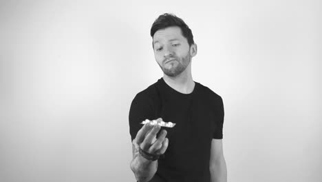 Grayscale footage of a tattooed man with short hair and a trimmed beard lifting up white medical pills or tablets in a package, looking at them and shaking his head, showing that he is not happy.