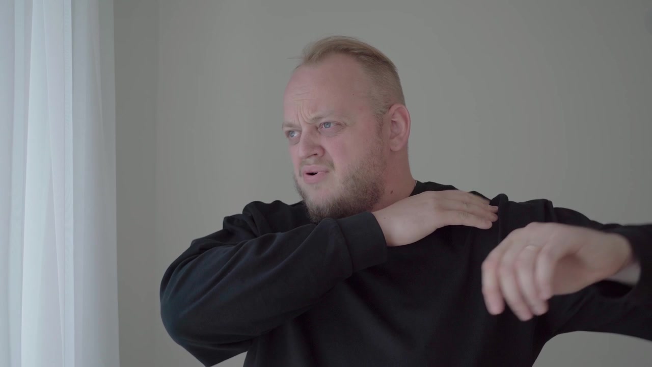 Middle-aged man in a dark sweatshirt winces and reaches across his body to grab his sore shoulder indoors.