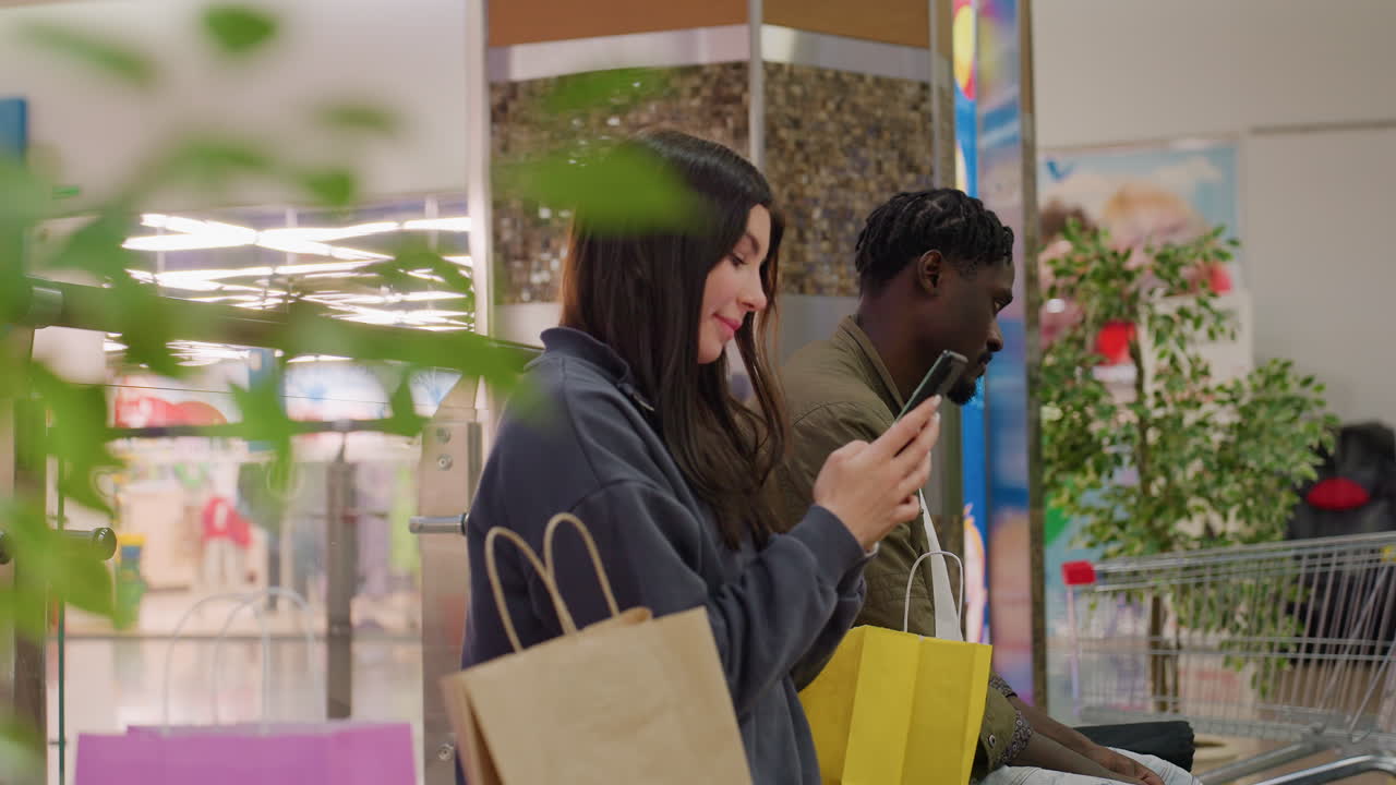 Smiling woman holding smartphone and shopping bags standing beside man also using phone while seated near shopping cart in indoor mall with greenery and bright background lights
