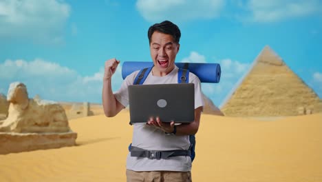 asian male hiker with mountaineering backpack looking at a laptop and screaming goal celebrating the success while traveling in pyramid of giza