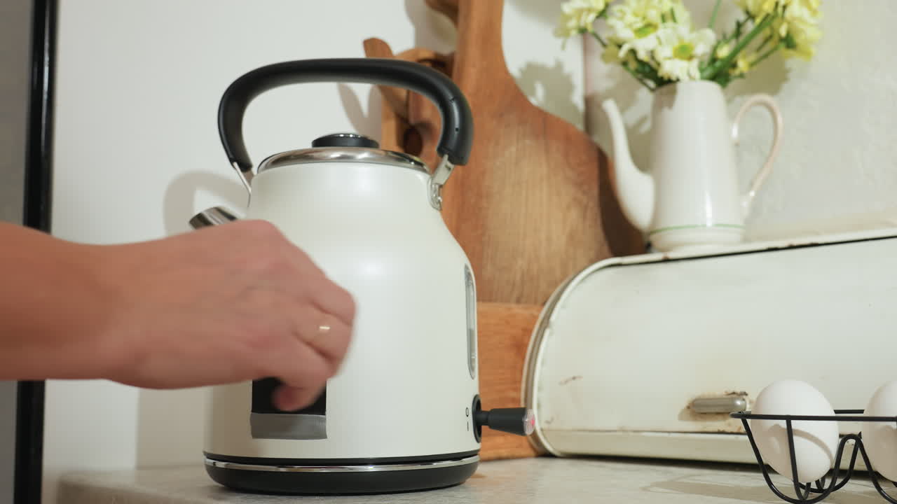 Electric kettle heating on kitchen countertop beside fresh eggs and vintage bread box with wooden chopping boards and ceramic jug with yellow flowers in cozy home interior under natural light