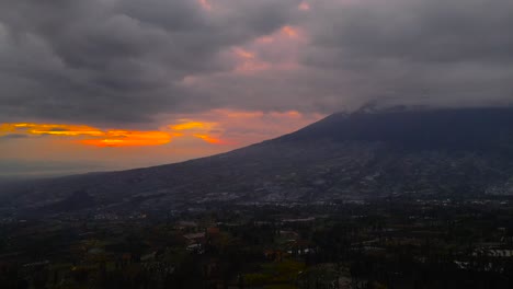 Vista-Aérea-Del-Paisaje-Rural-Con-Una-Enorme-Montaña-Y-Hora-Del-Amanecer-En-El-Cielo-Nublado---El-Cielo-Es-Naranja