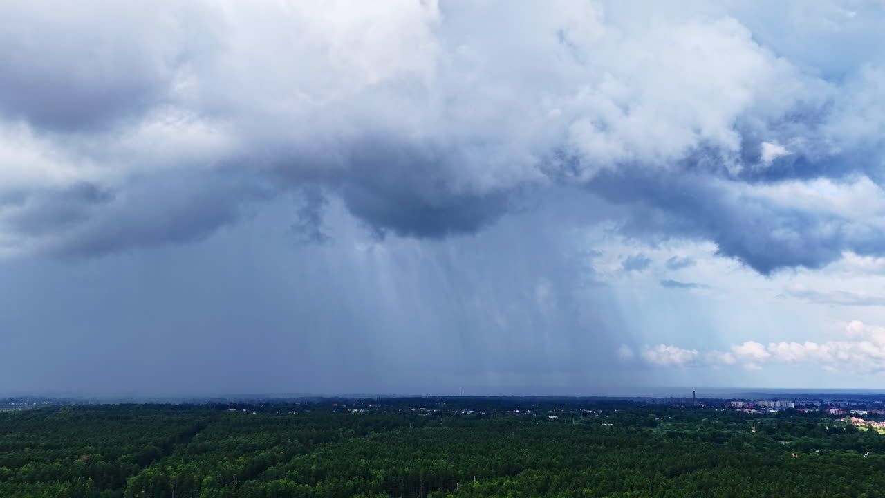 Heavy rain pours from dark dramatic clouds over a lush green forest landscape near Jelgava, Latvia, capturing the power and beauty of summer storms